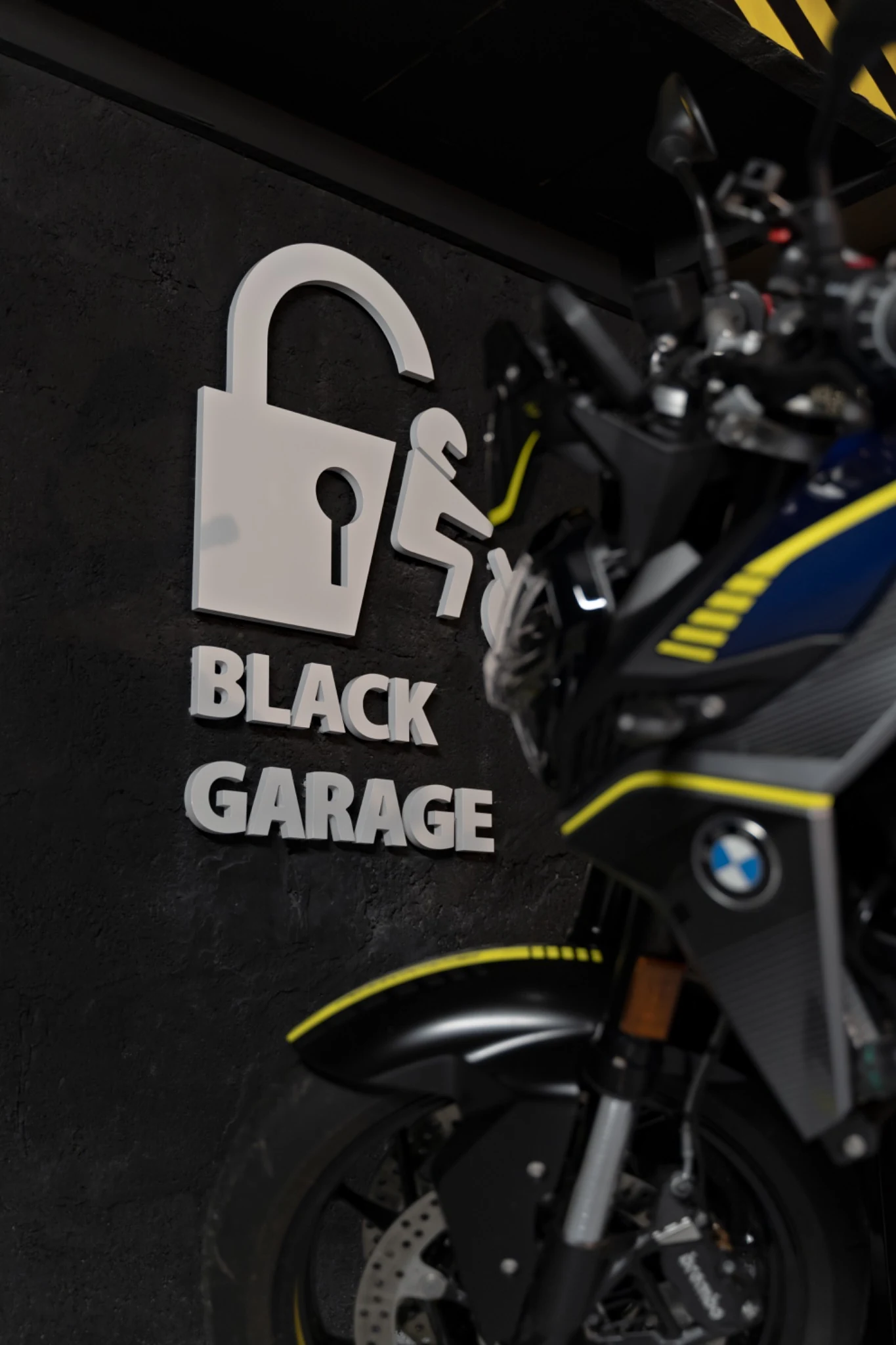 Accueil 75 Logo Black Garage sur un mur sombre, à côté d’une moto noire et jaune stationnée à l’intérieur.