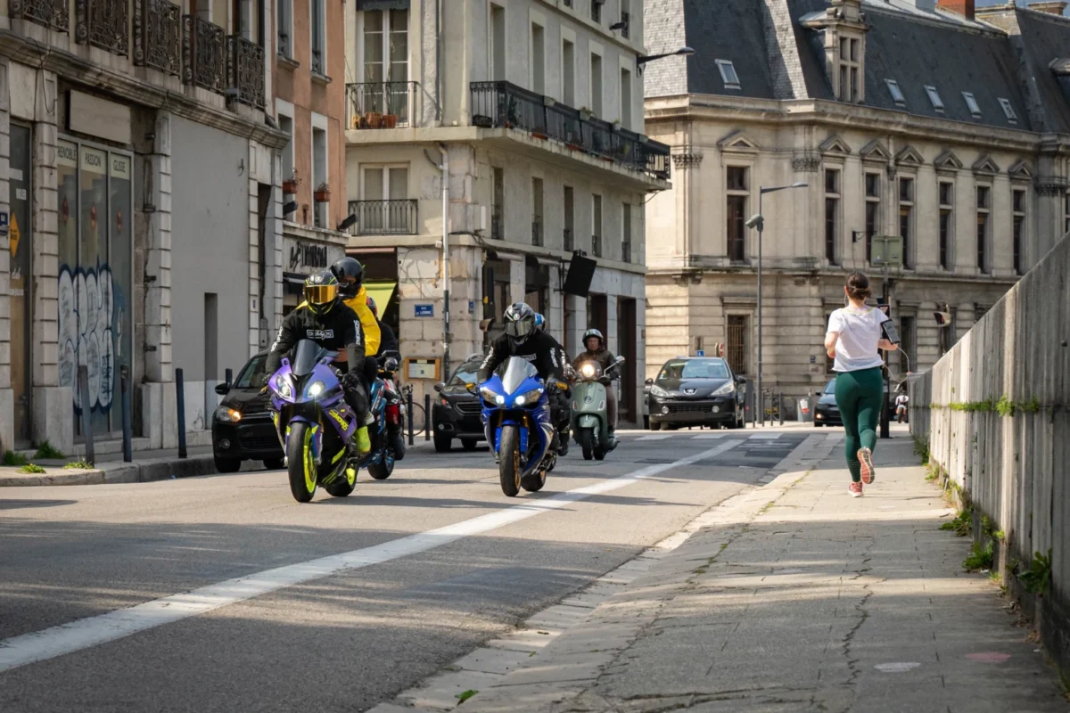 Plusieurs motards et un scooter circulant dans une rue urbaine bordée d’immeubles.