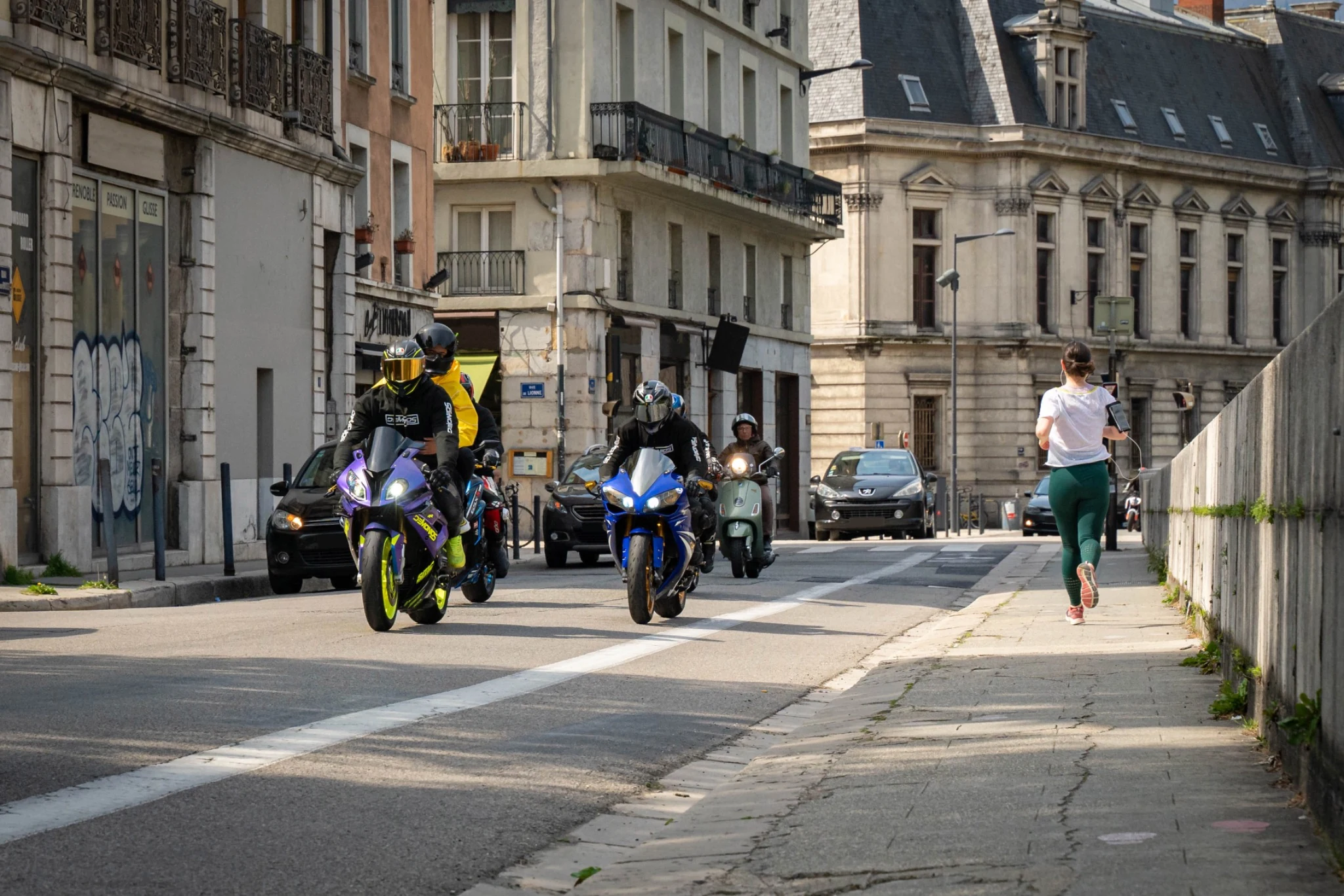 Accueil 73 Plusieurs motards et un scooter circulant dans une rue urbaine bordée d’immeubles.